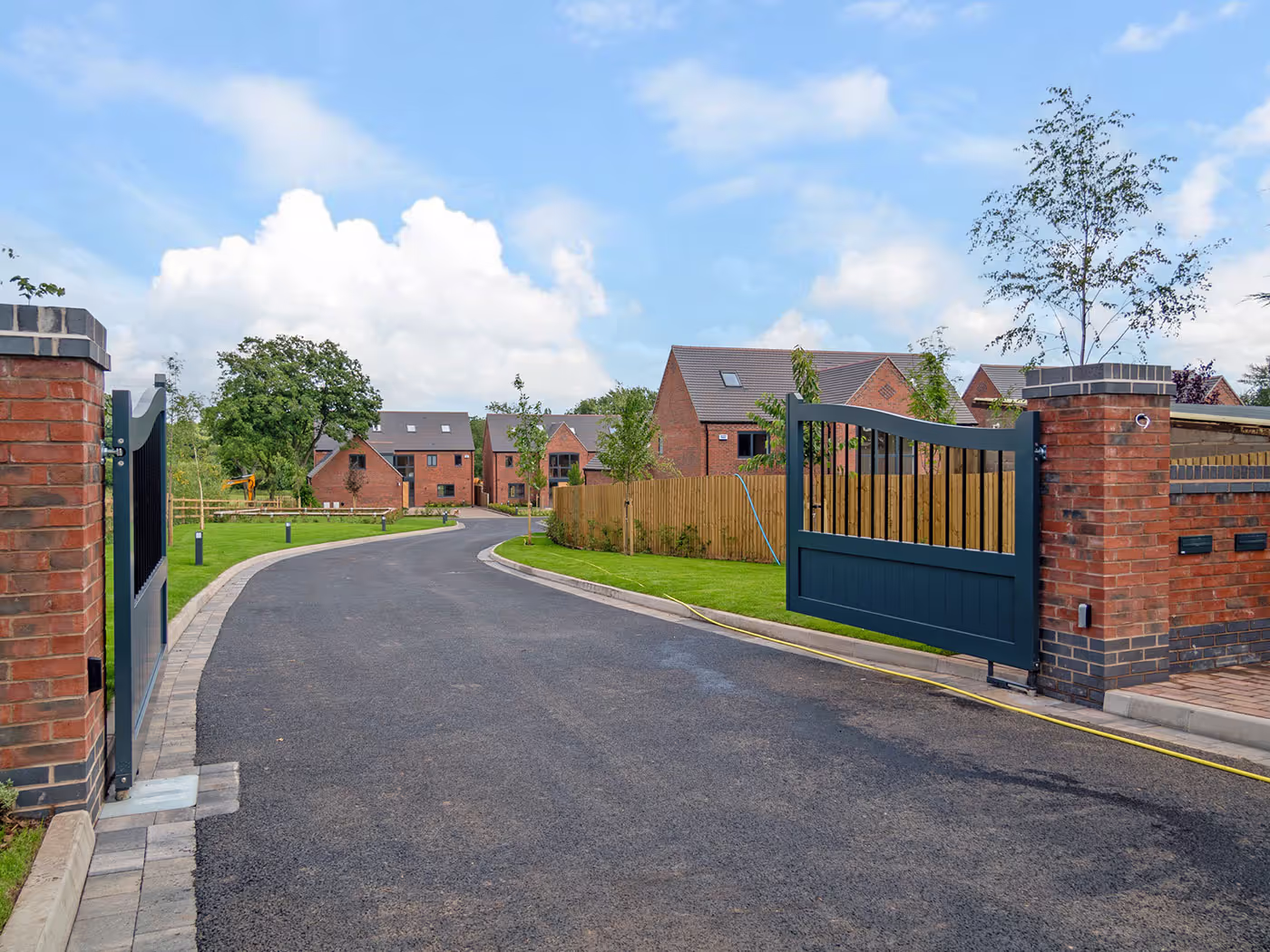 Residential street view with open black metal gates, brick pillars, and red brick houses under a blue sky.