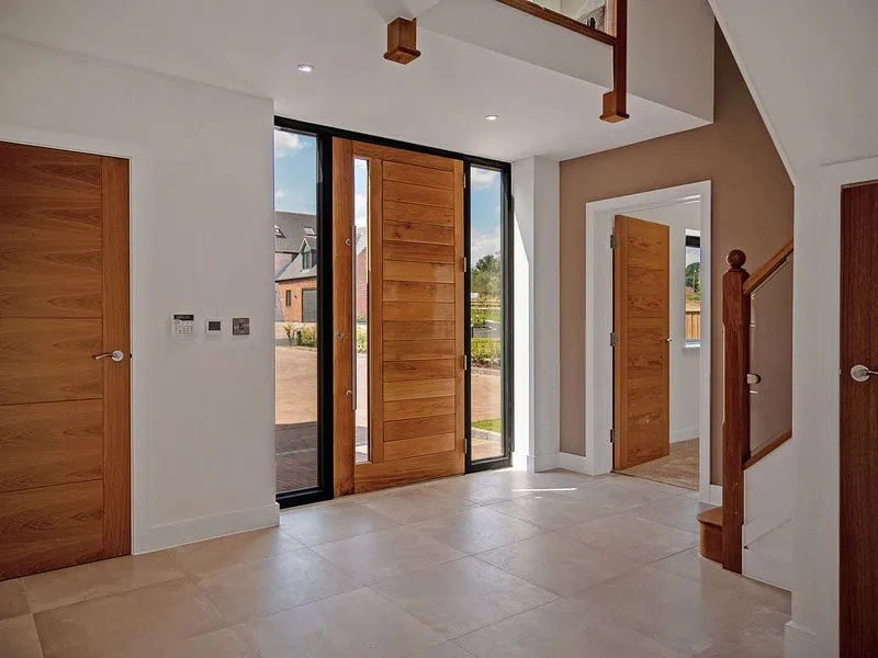 Modern home's entryway with a large wooden front door, glass panels, tiled floor, and wooden staircase.