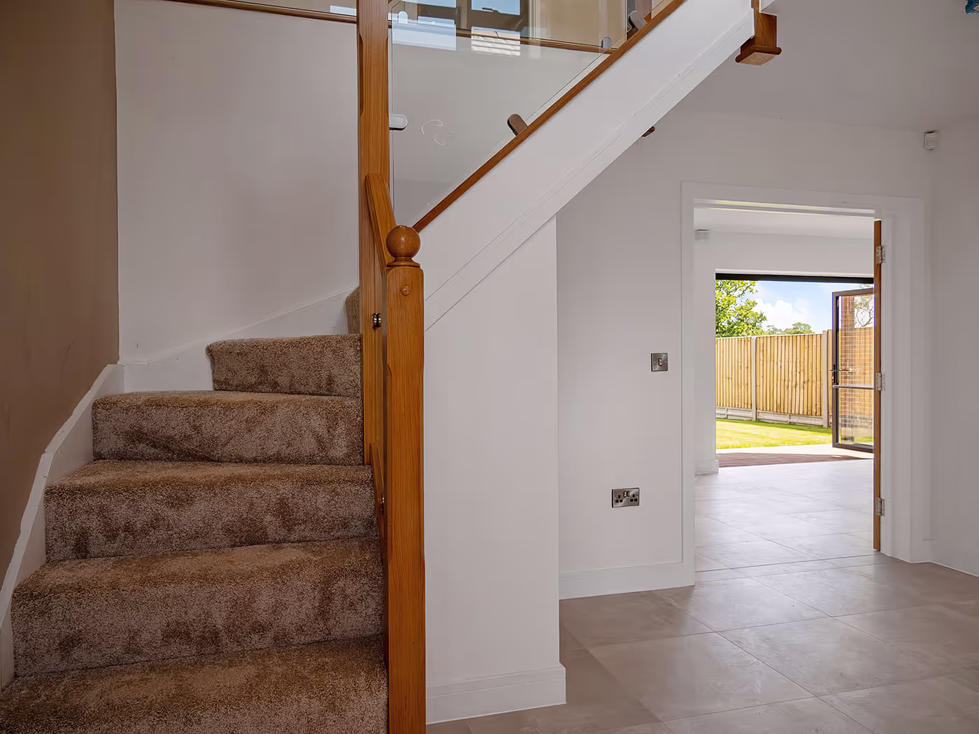 Carpeted staircase with wooden banister beside open doorway revealing tiled floor and garden outside.