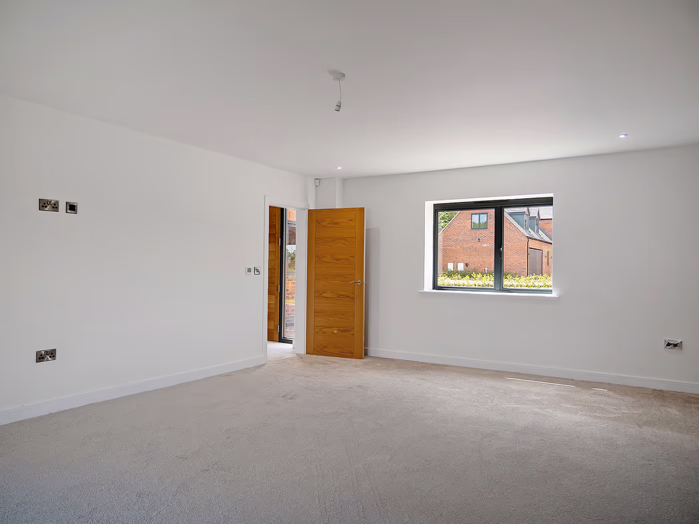Empty room with beige carpet, white walls, wooden door open, and window looking outside brick building.