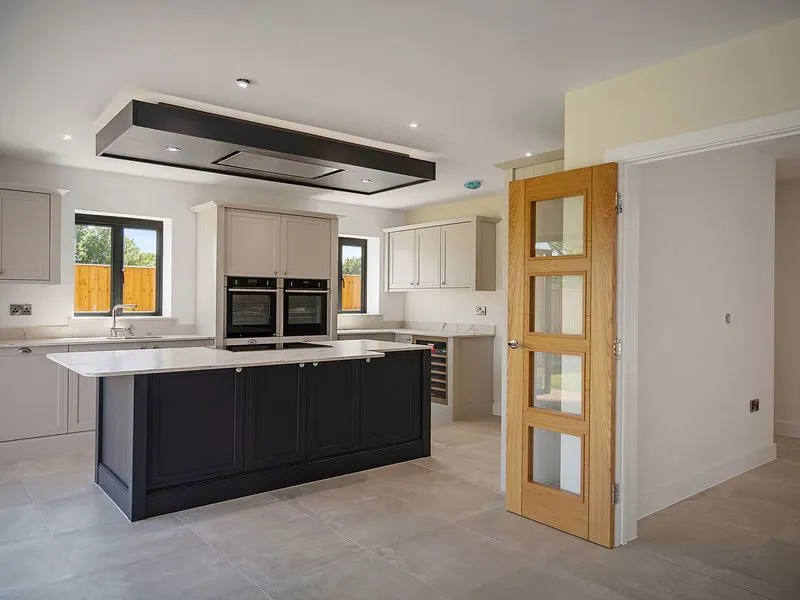 Modern kitchen with black island, beige cabinets, double ovens, and a wooden door with glass panels.