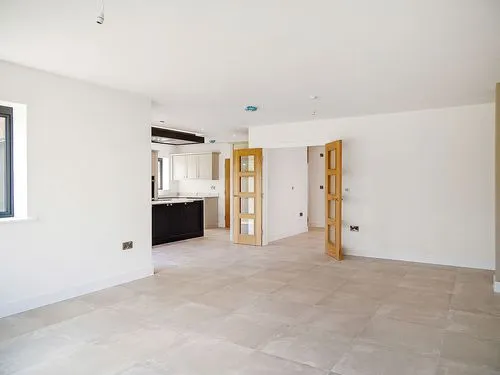 Empty open-plan room with beige tiled floor, white walls, wooden door, and kitchen area in the back.