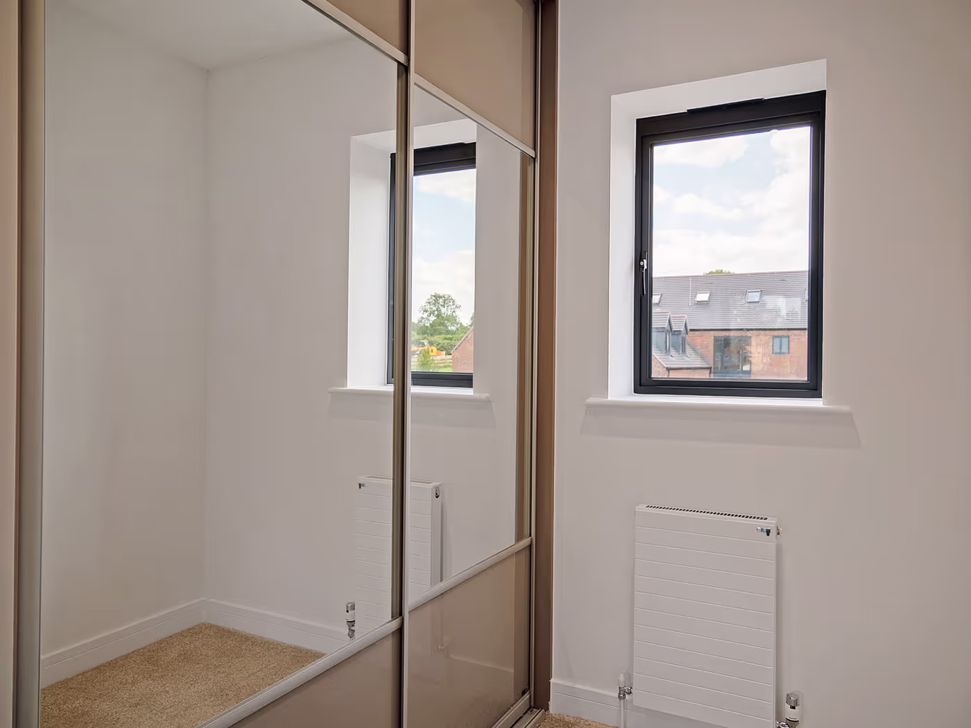 Small room with beige carpet, white walls, radiator, window, and mirrored sliding closet doors.