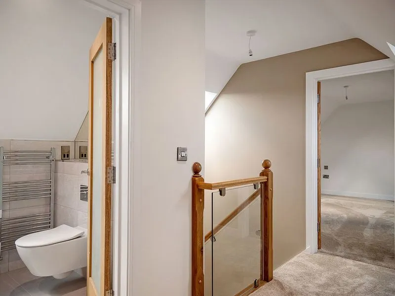 Interior hallway with beige walls, carpeted floor, wooden staircase railing, and a bathroom with a wall-mounted toilet.