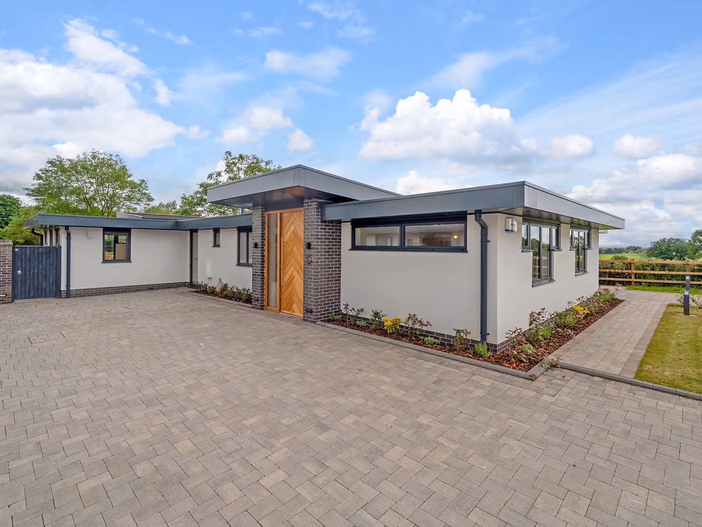 Modern single-story house with flat roofs, wooden front door, large driveway, and garden beds.