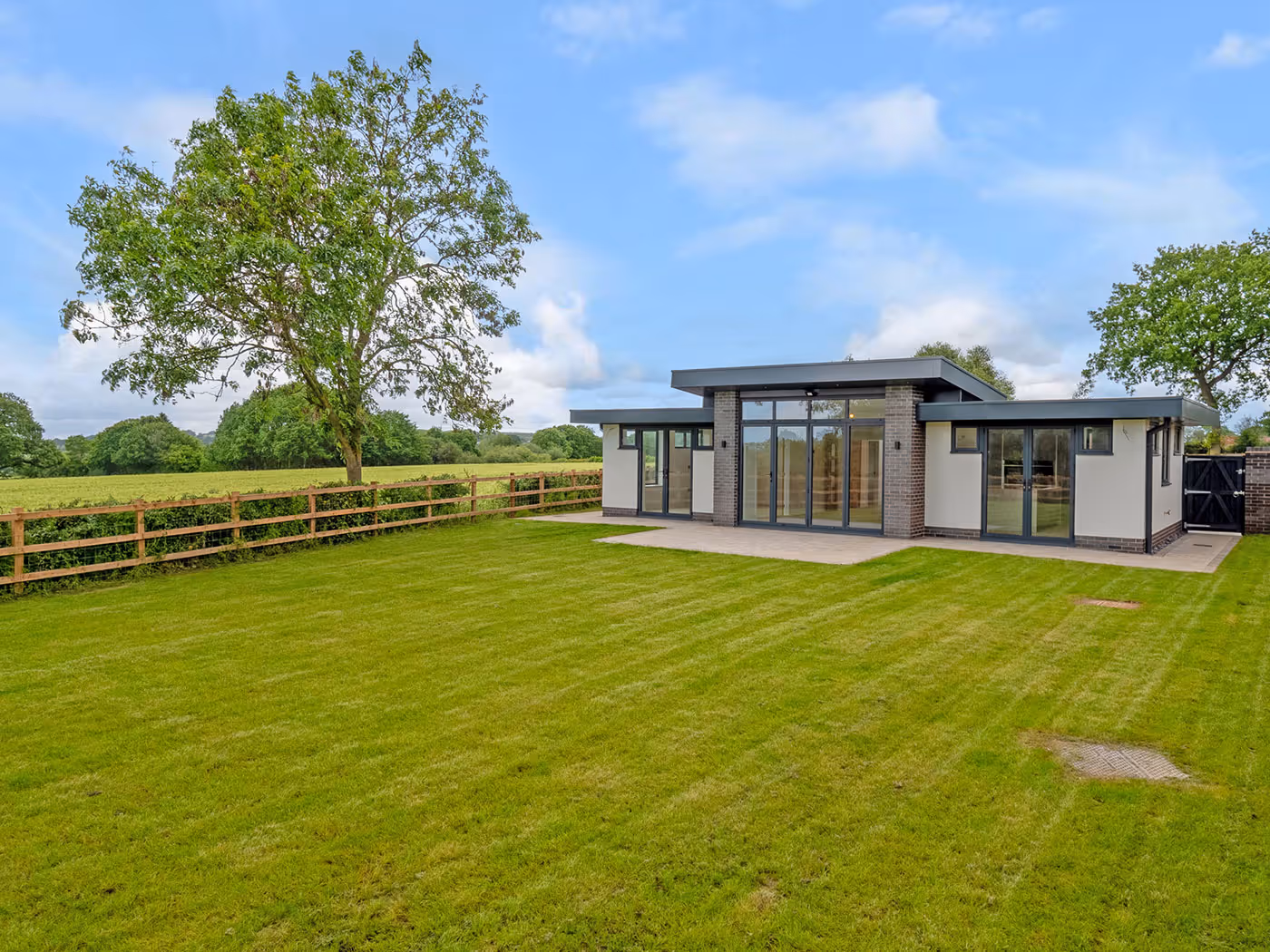 Modern single-story house with large glass doors and windows, green lawn, wooden fence, and trees under blue sky.