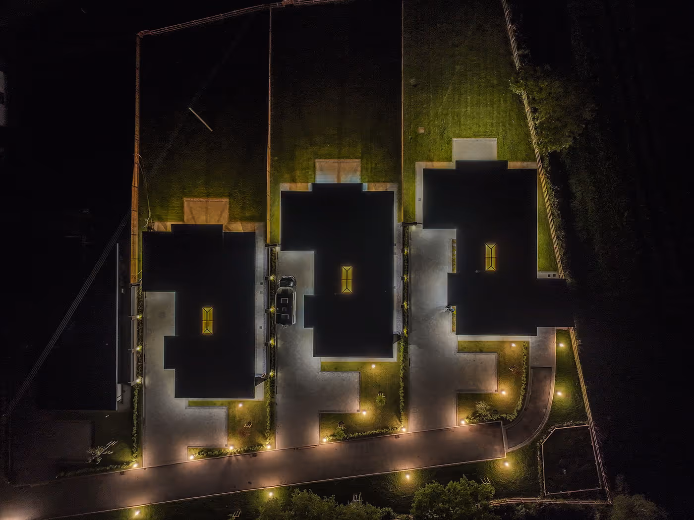Aerial night view of three houses with lit driveways, yards, and a street in front.