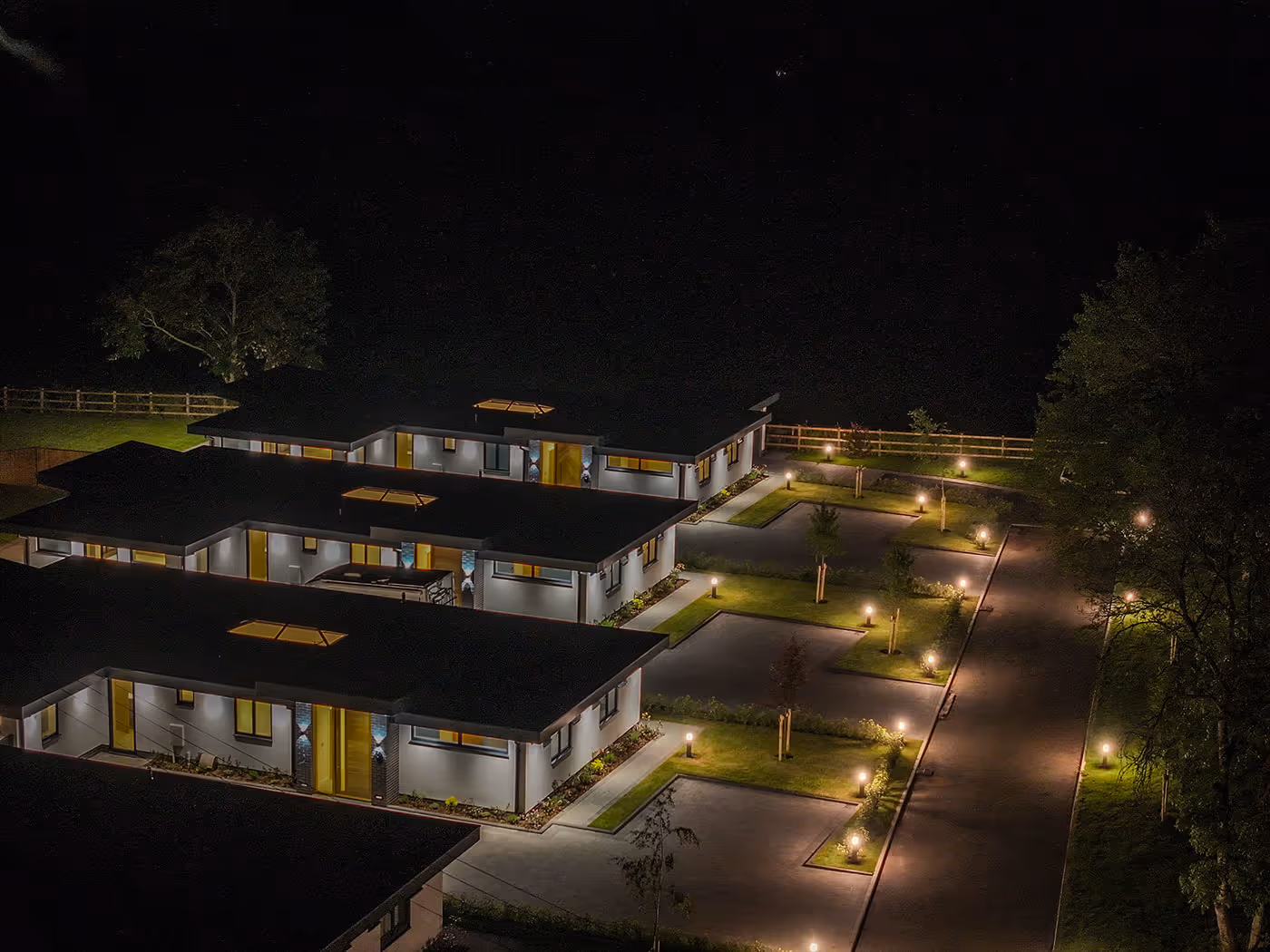 Night aerial view of illuminated modern houses with driveways and landscaped lawns.
