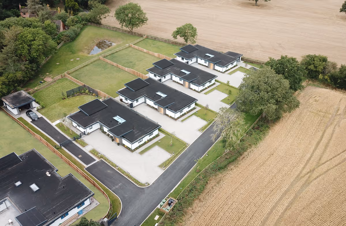 Aerial view of modern single-story houses with black roofs and green lawns beside a harvested field.