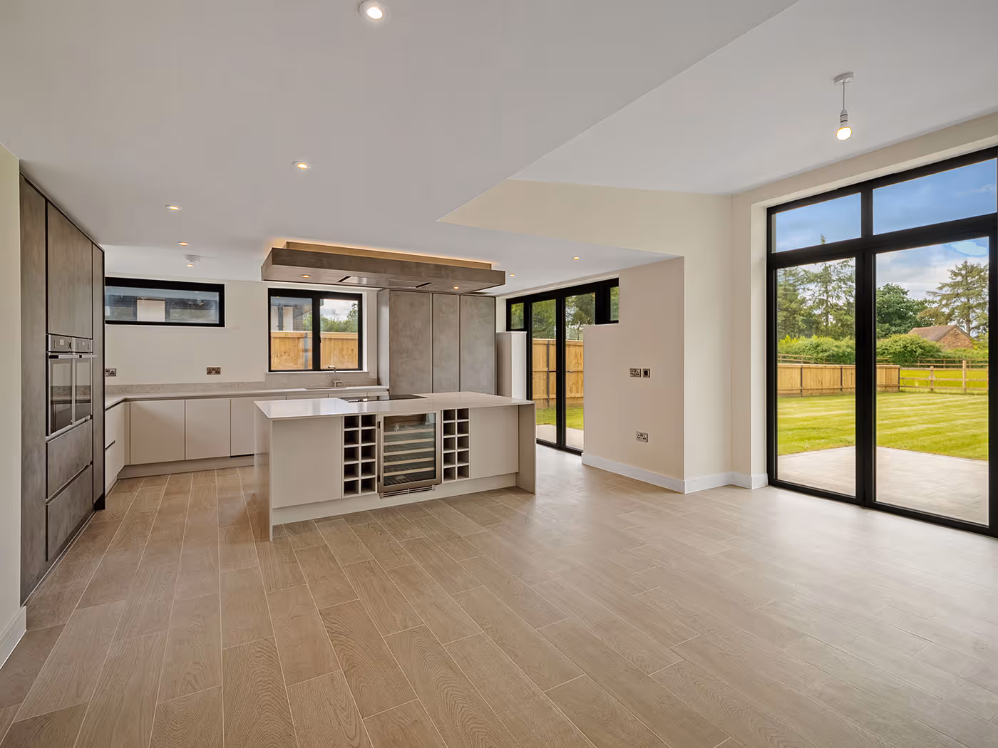 Modern kitchen with island, wine cooler, light wood floor, large windows, and glass doors to a green yard.