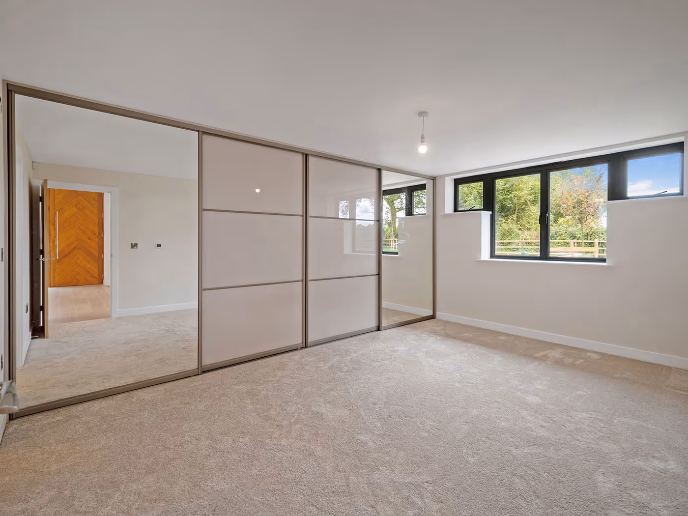 Empty bedroom with beige carpet, large sliding wardrobe with mirrors, and windows overlooking greenery.