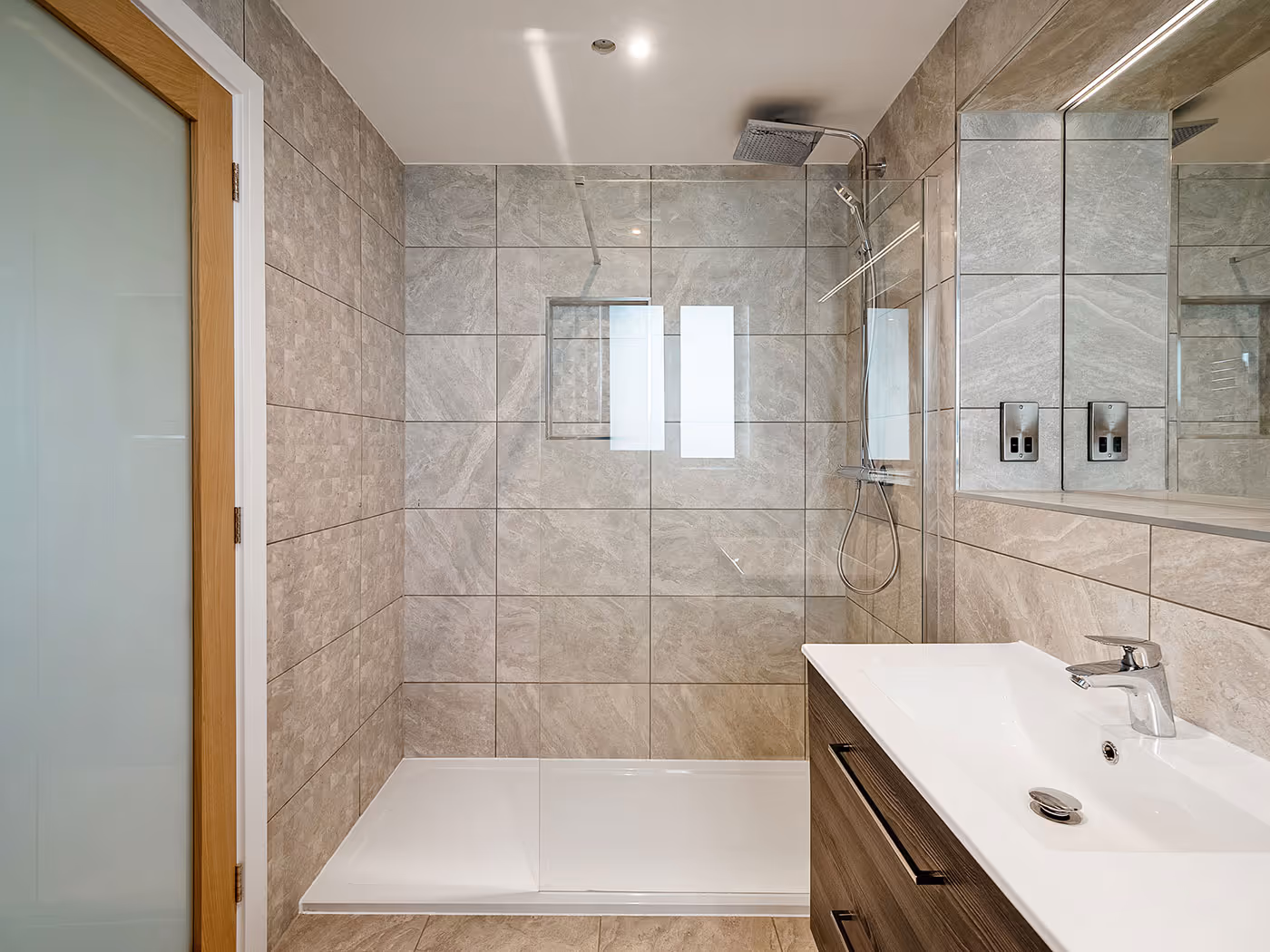 Modern bathroom with walk-in shower, beige tiled walls, glass partition, and wooden vanity with white sink.
