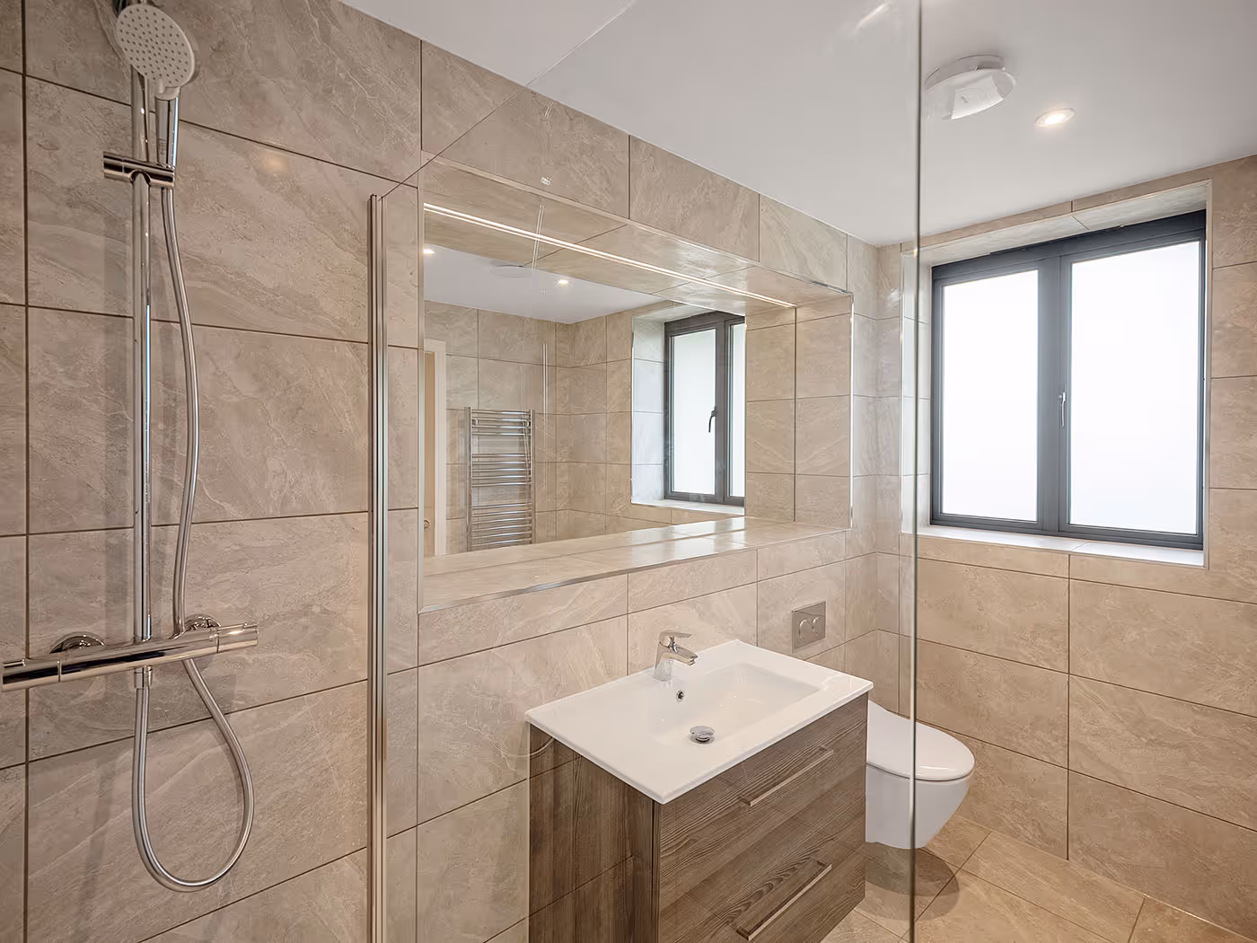 Modern bathroom with beige tiles, wooden vanity, wall-mounted toilet, large mirror, and window.