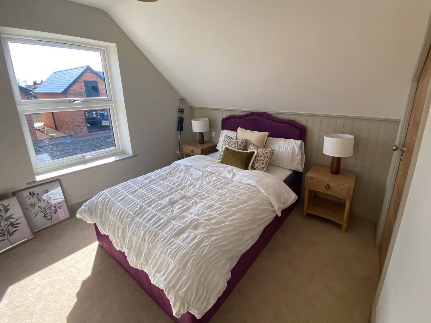 Cozy bedroom with slanted ceiling, purple bed, white bedding, wooden nightstands, and lamps near window.