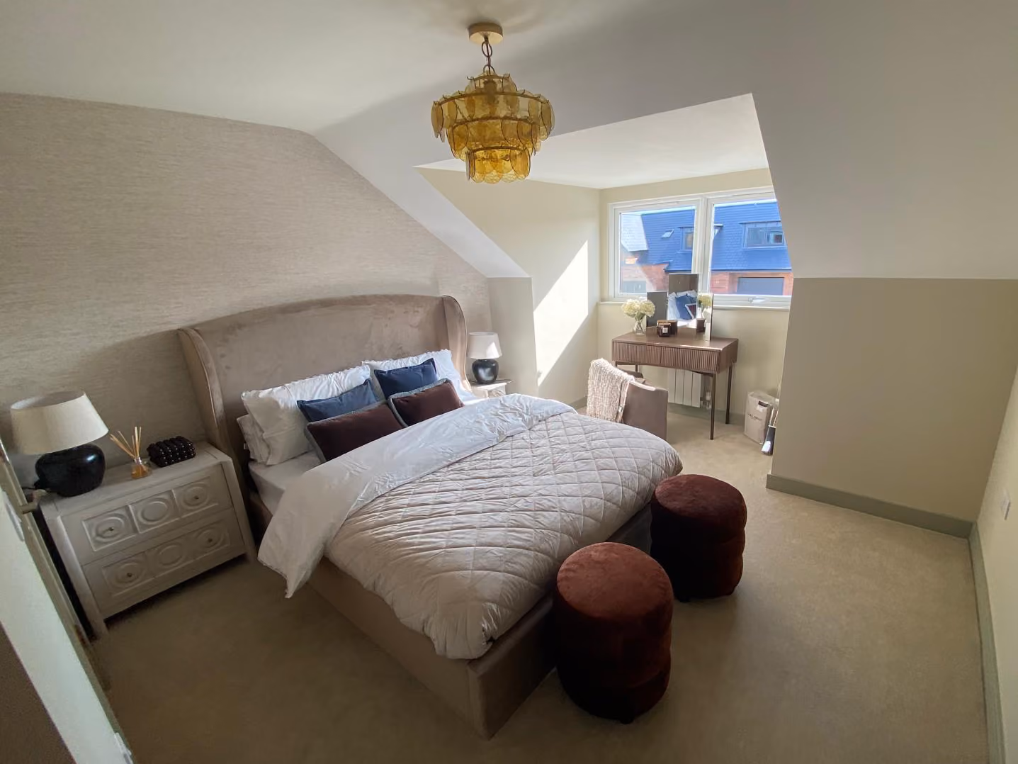 Bedroom with a large bed, beige walls, two bedside tables, a window, and two red ottomans at the foot.
