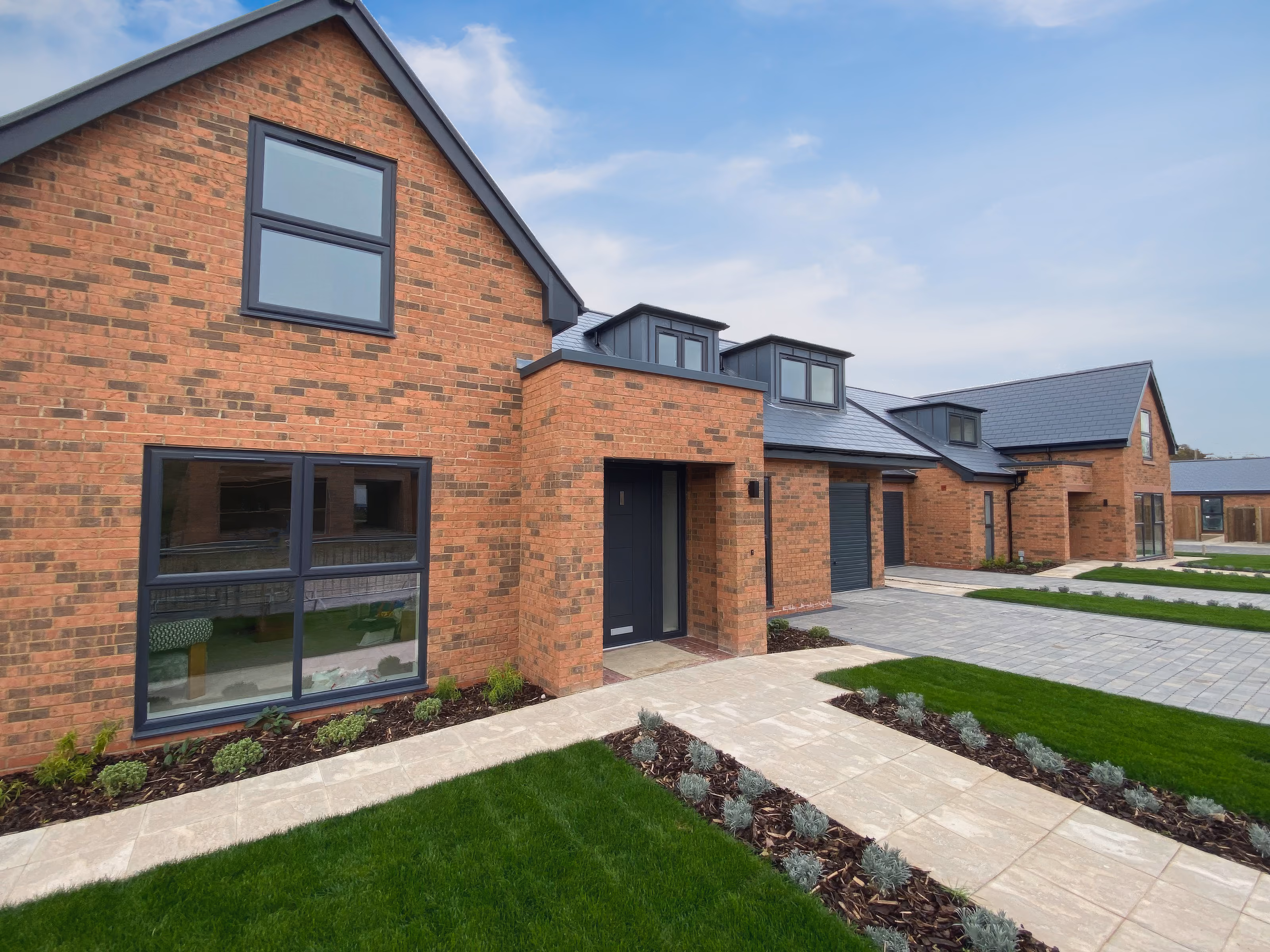 Modern brick homes with large windows, hanging flower baskets, and a green lawn under a cloudy sky.