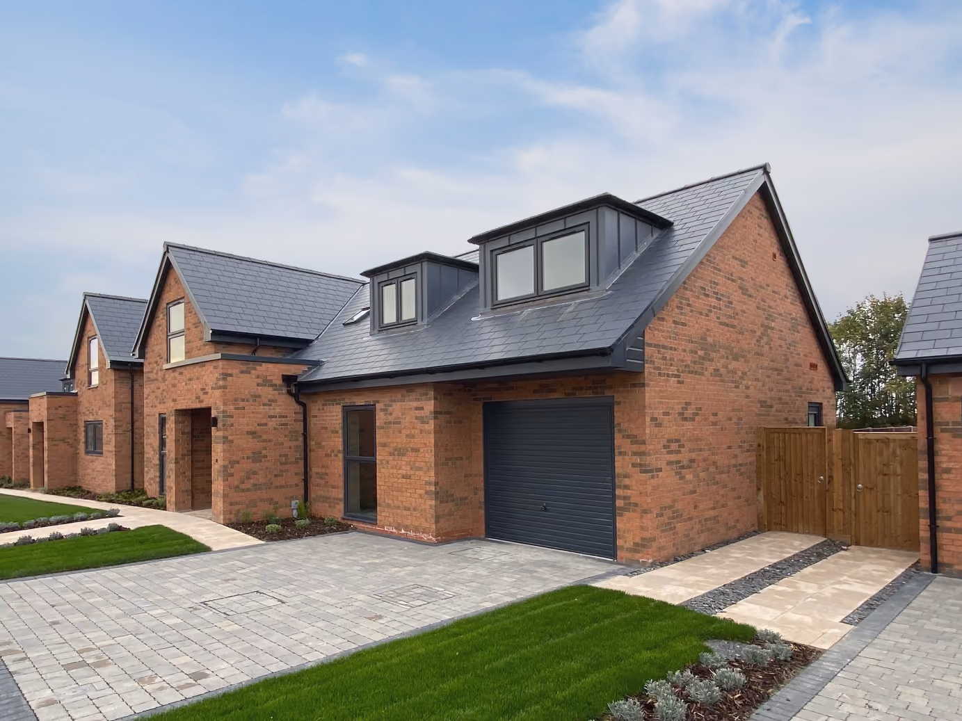 Row of modern brick houses with dark gray roofs, garages, driveways, and green lawns under a clear sky.
