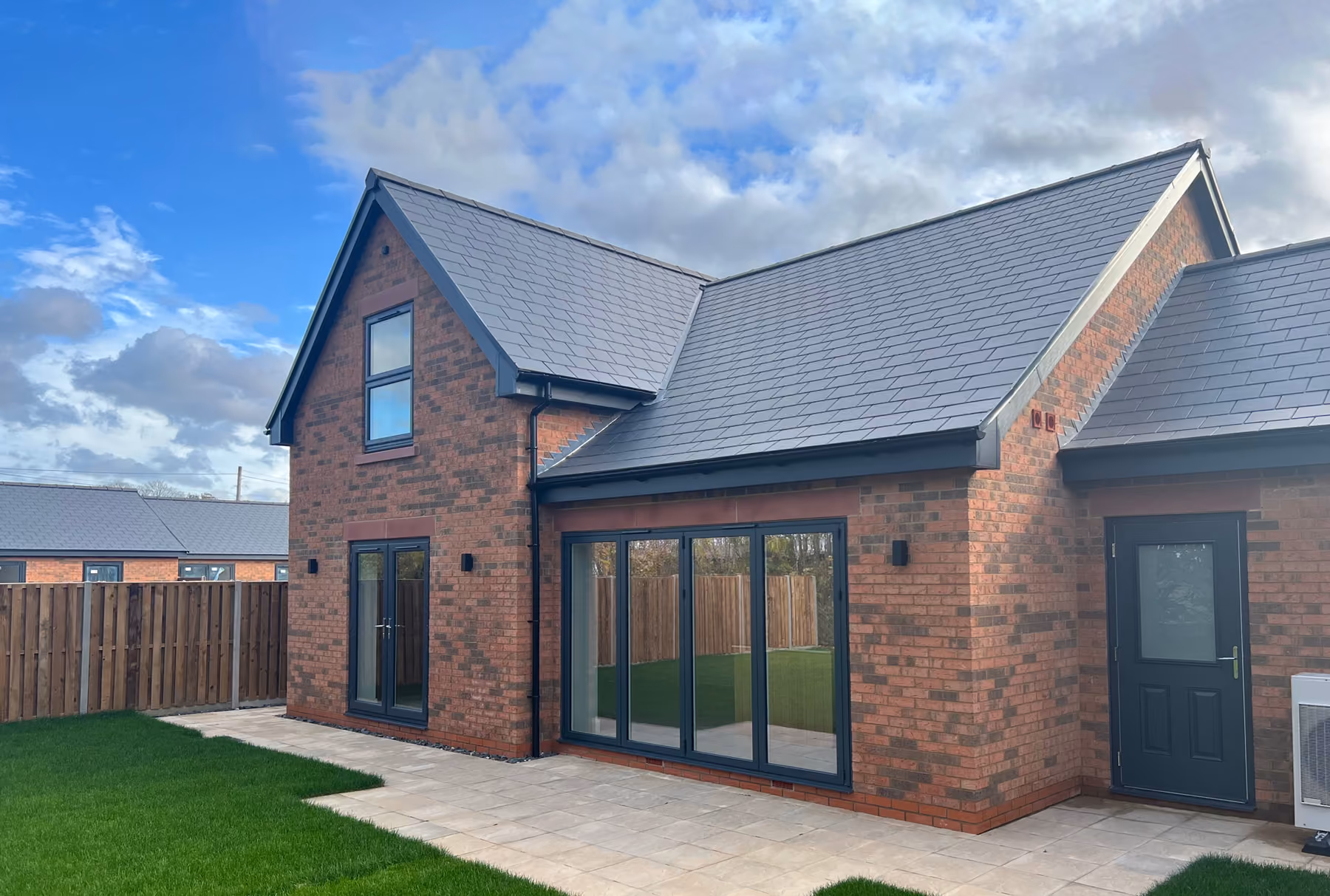 Modern red brick house with grey roof, large windows, patio, and green lawn under cloudy sky.