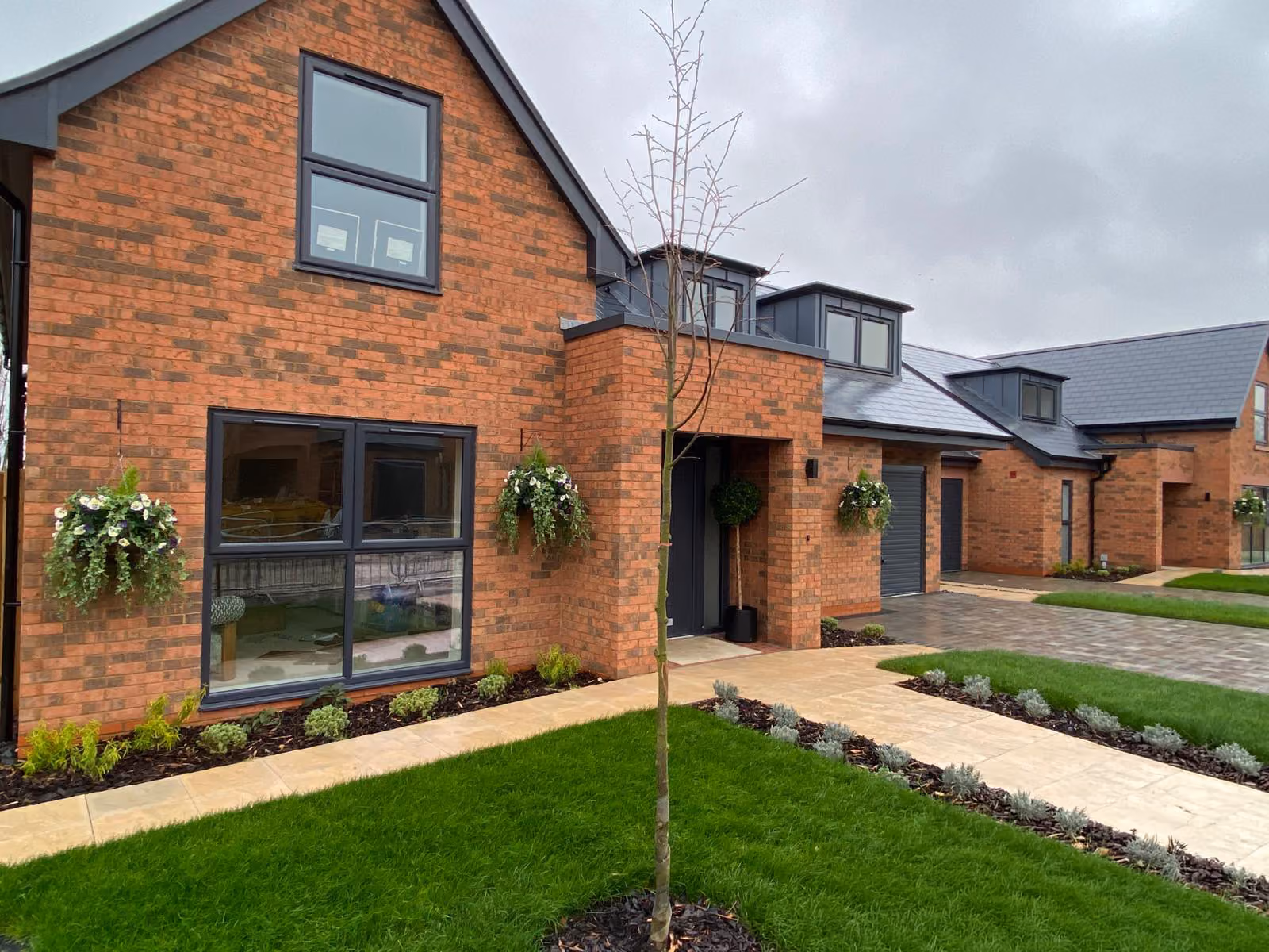 Modern brick homes with large windows, hanging flower baskets, and a green lawn under a cloudy sky.