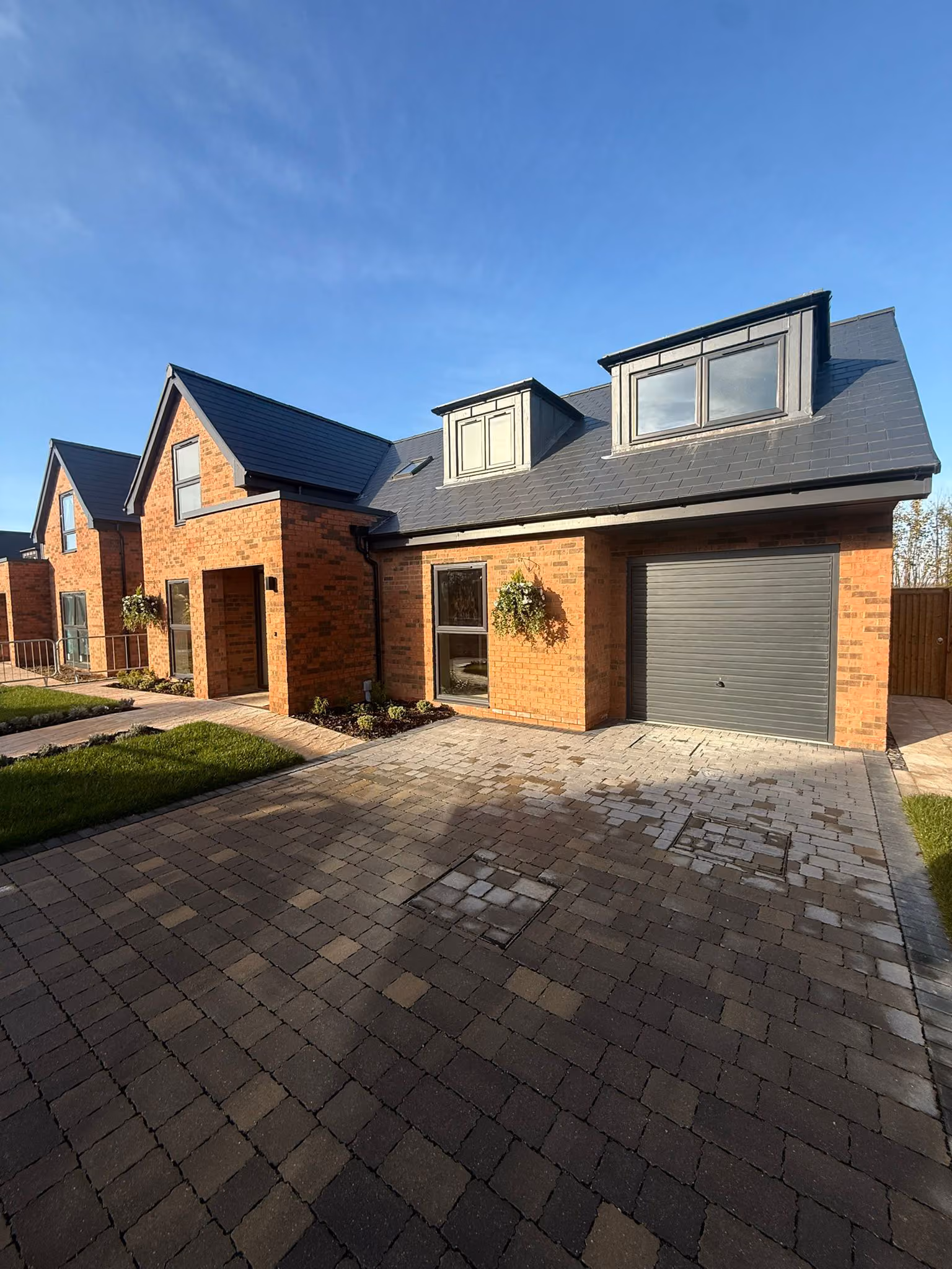 Brick house with slate roof, two dormer windows, garage, paved driveway, and hanging plants.
