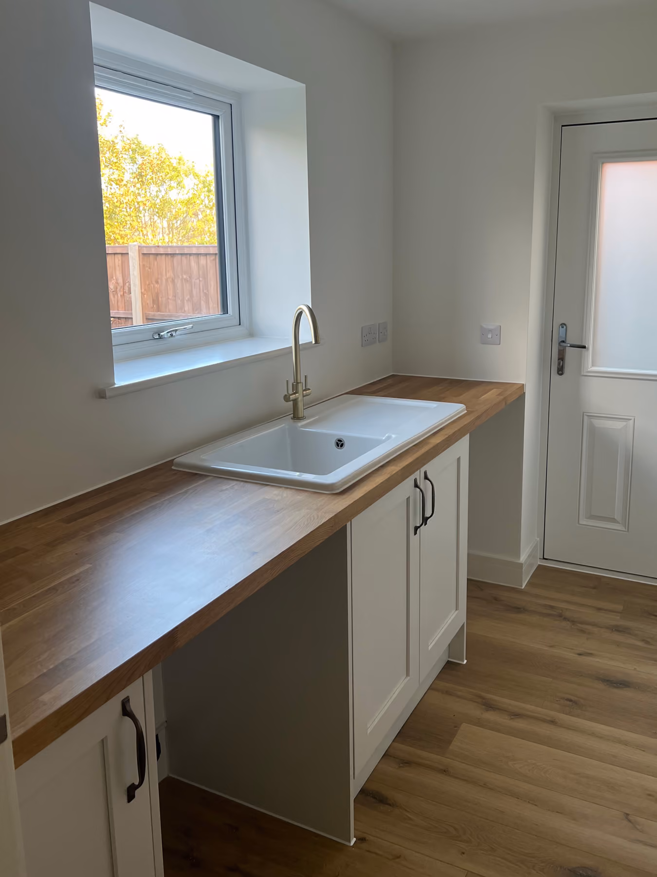 Clean kitchen area with wooden countertop, white sink, brass faucet, white cabinets, window, and door.