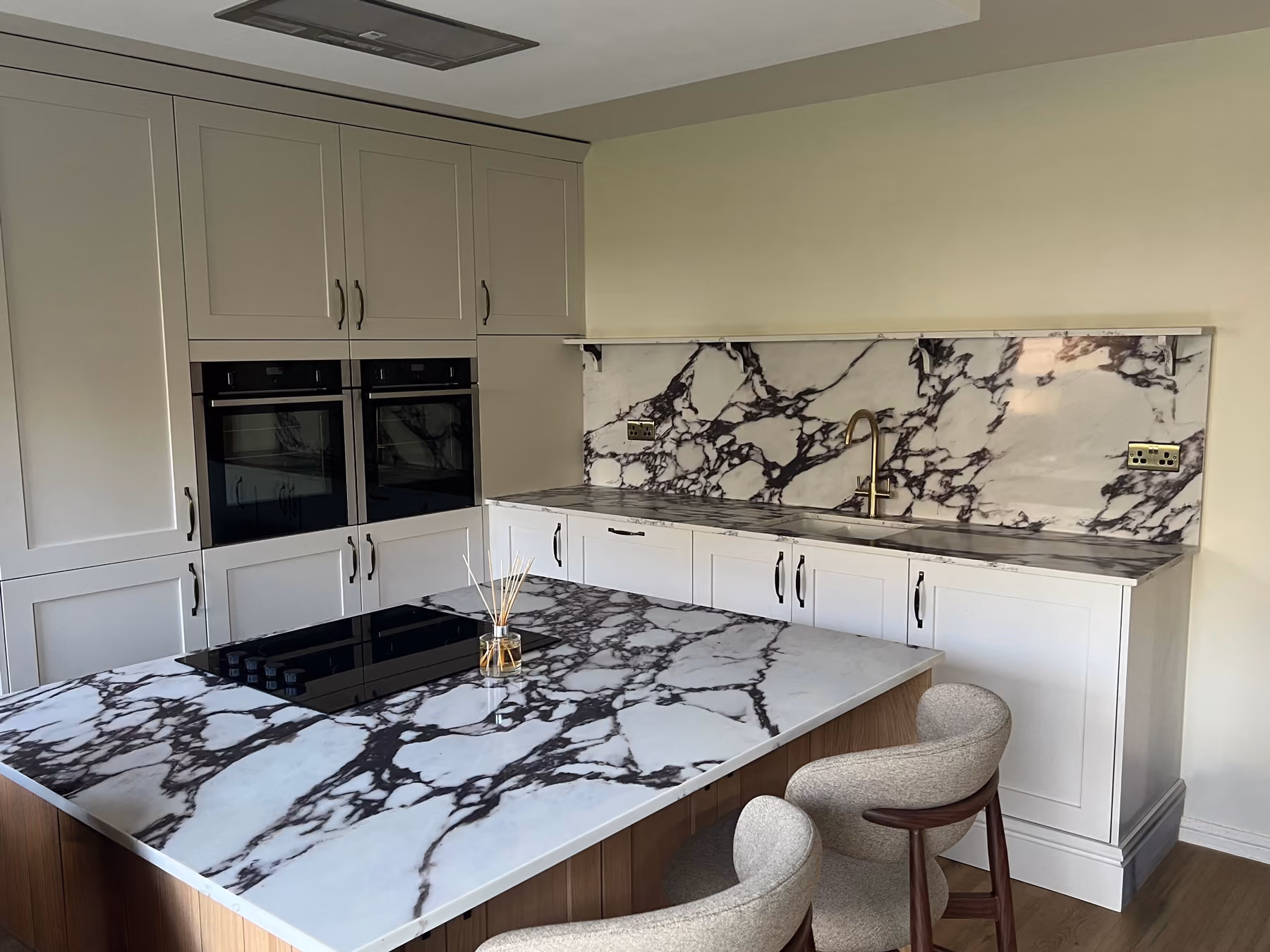 Modern kitchen with white cabinets, marble countertops, island with cooktop, and two gray chairs.