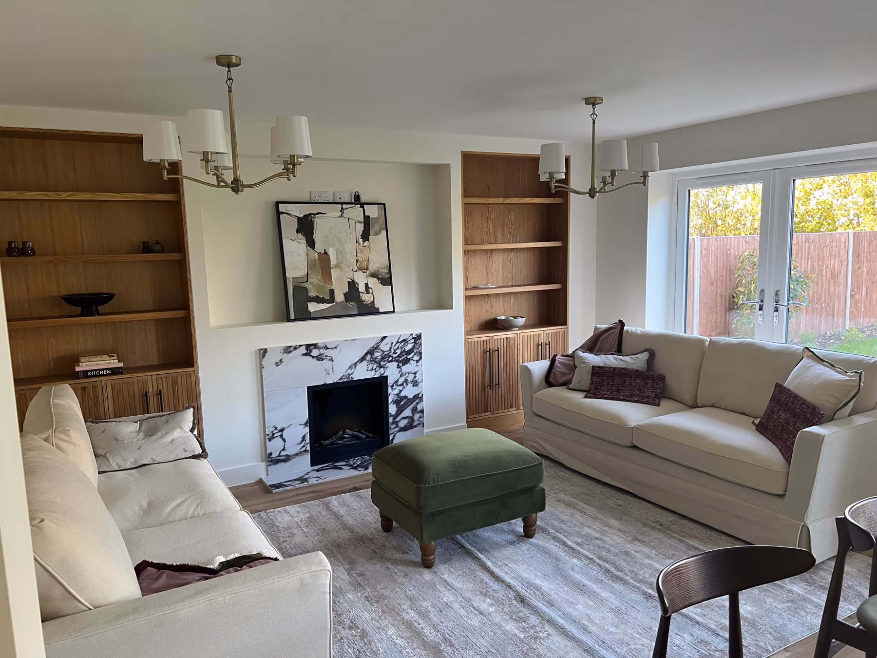 Living room with two beige sofas, green ottoman, built-in wooden shelves, marble fireplace, and glass doors.