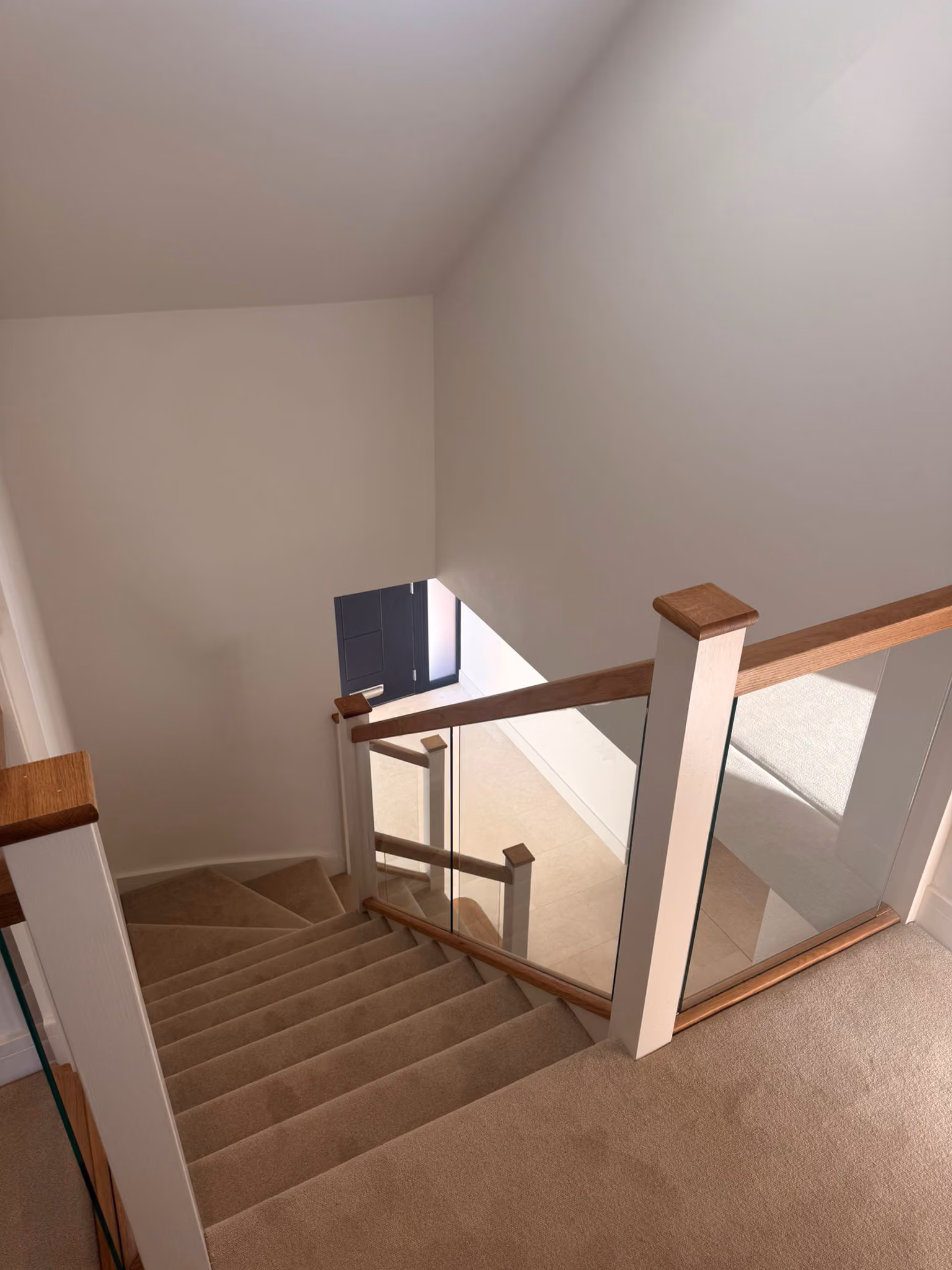Carpeted staircase with wooden and glass railings leading down to a dark entry door.