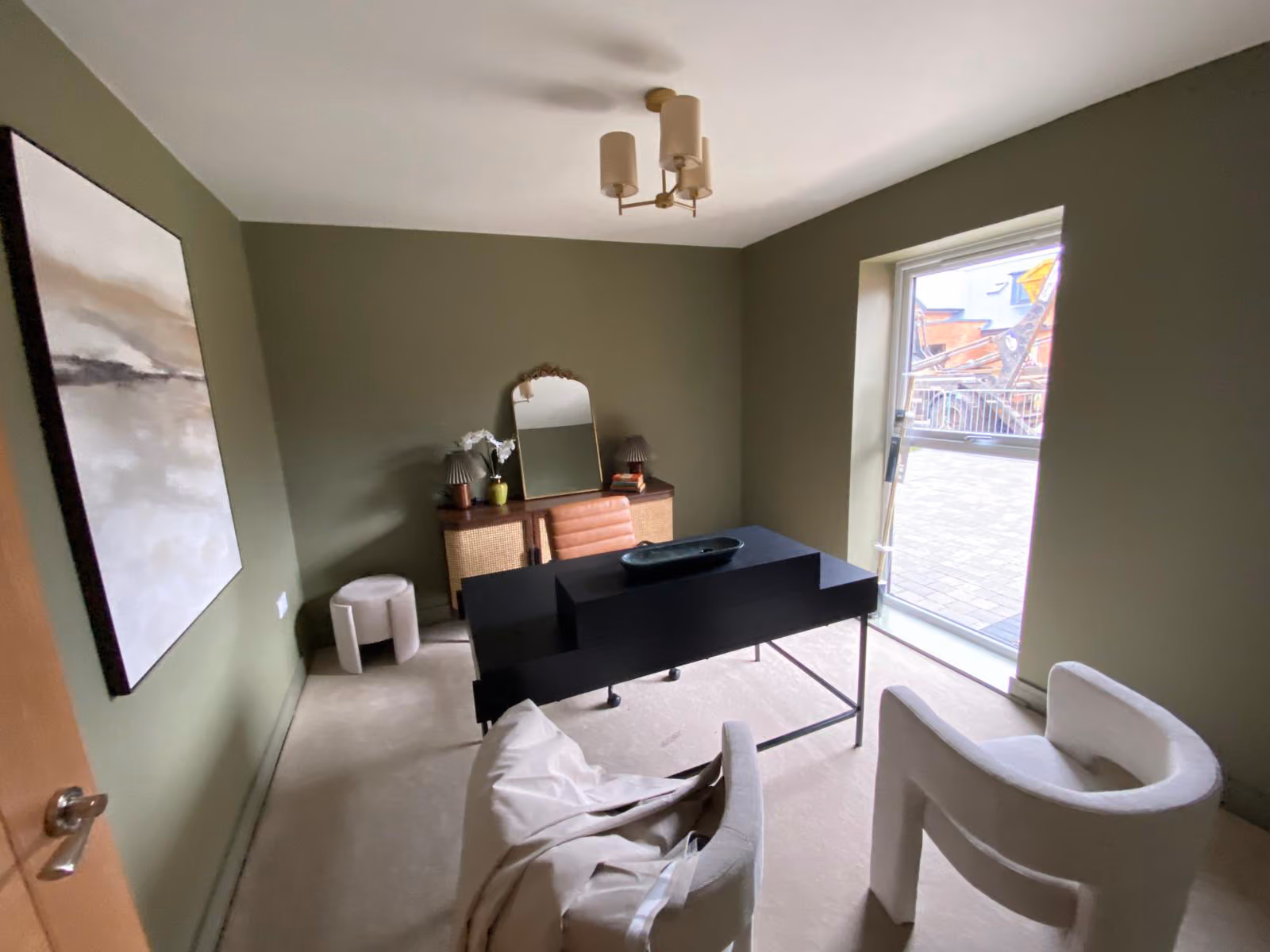 Modern office with olive walls, black desk, two white chairs, abstract painting, and window with view.