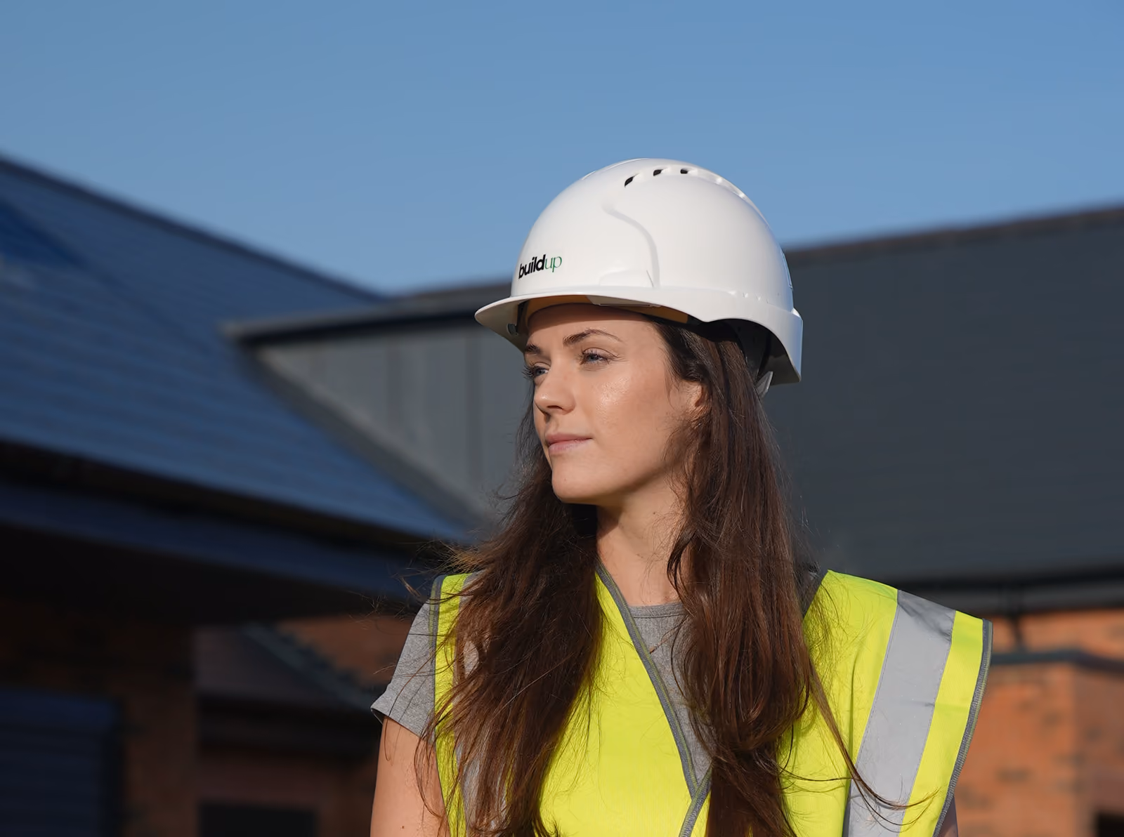 Rosey Cassidy in a white hard hat and yellow reflective vest standing outdoors against a building and clear blue sky.
