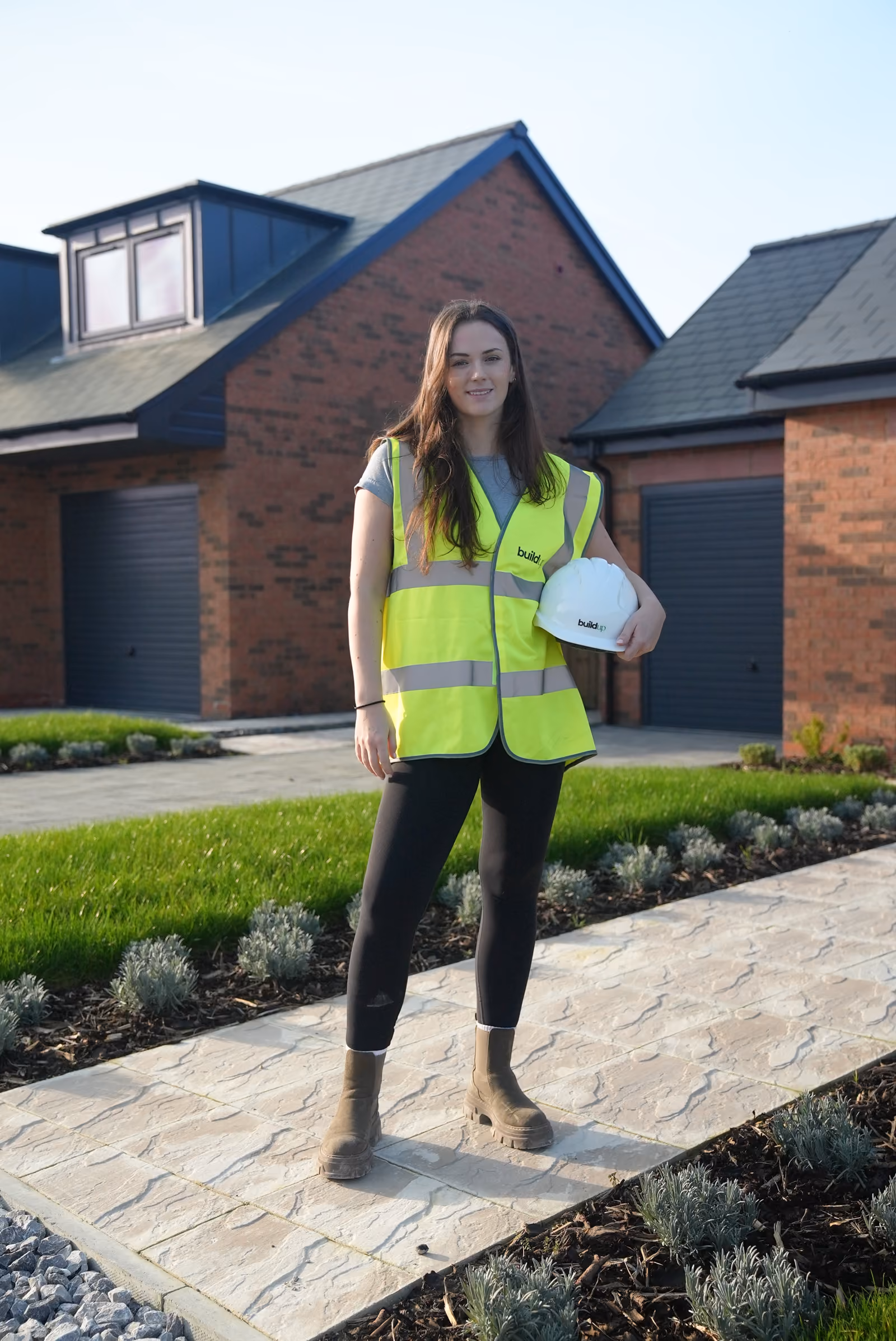 Rosey Cassidy in a yellow safety vest holding a white hard hat standing on a stone path near brick houses.