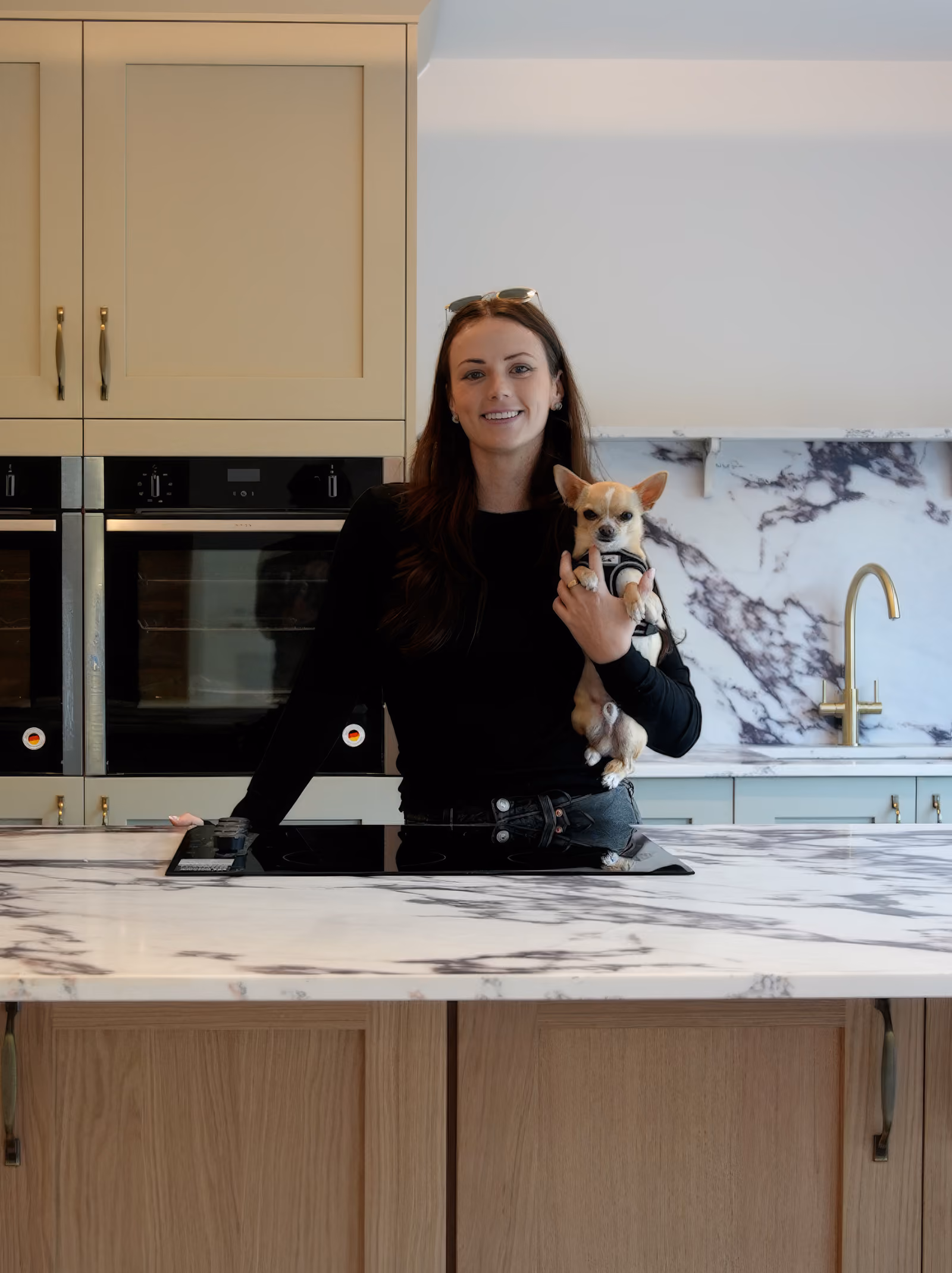 Rosey Cassidy in black shirt holding a small dog in a modern kitchen with marble countertops and gold faucet.